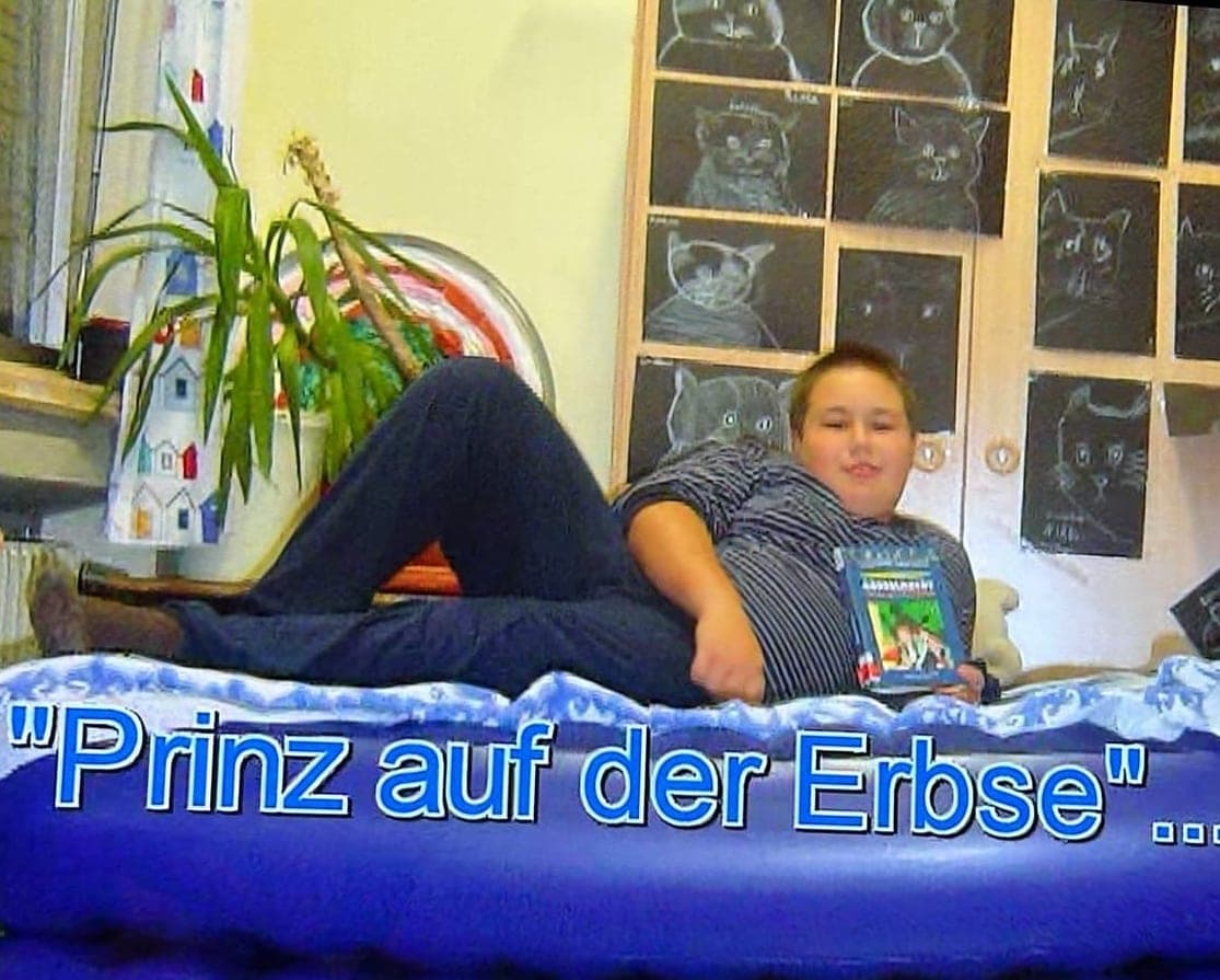 Fernsehaufnahme aus Dennis' Kindheit: er liegt als Junge auf einer Luftmatratze, TV-Einblendung „Prinz auf der Erbse“