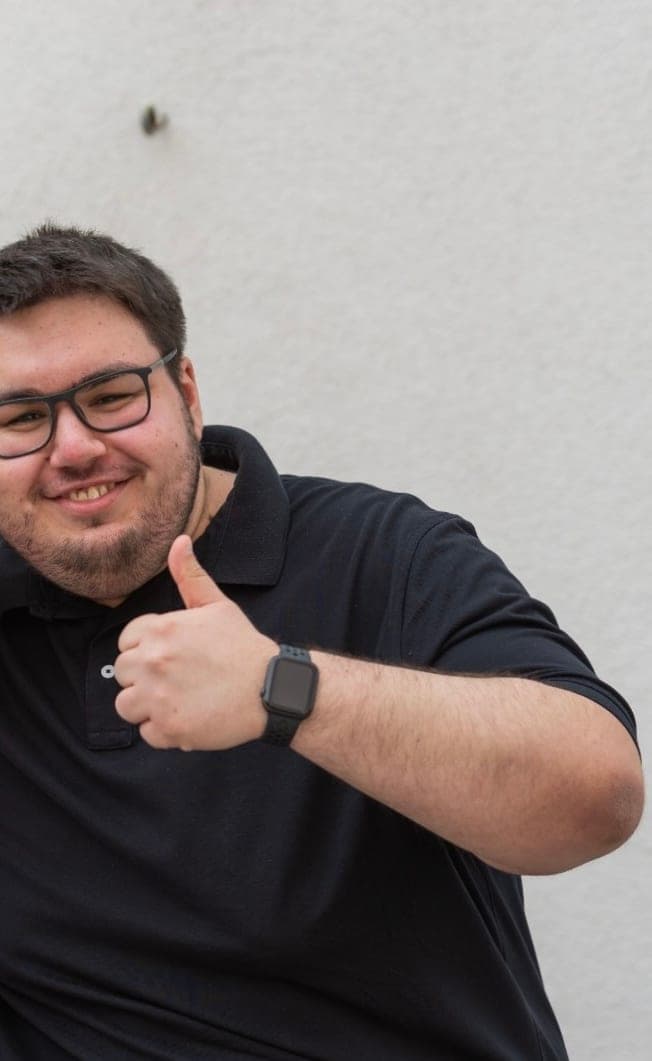 Dennis im dunklen Polohemd vor einer hellen Wand, lächelnd mit Daumen nach oben